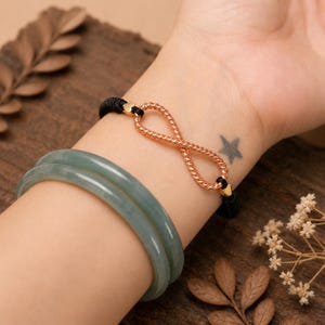 May include: A bracelet featuring a rose gold-toned infinity symbol made of twisted metal, attached to a black cord, paired with two smooth, translucent, pale green jade-colored bangle bracelets worn on the same wrist.