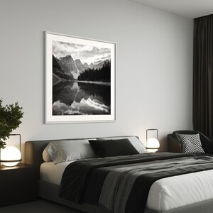 May include: A black and white photograph of a mountain lake with snow-capped peaks and a forest in the background. The photo is framed in a white frame and hangs on a white wall above a bed with black and white bedding.
