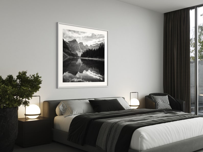 May include: A black and white photograph of a mountain lake with snow-capped peaks and a forest in the background. The photo is framed in a white frame and hangs on a white wall above a bed with black and white bedding.