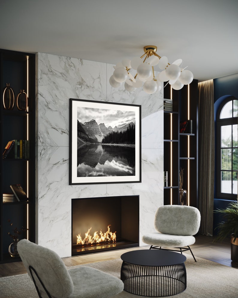 May include: A modern living room interior with a fireplace, two armchairs, a coffee table, and a black and white photograph of a mountain lake hanging on the wall. The room has a marble wall and a black and gold chandelier.