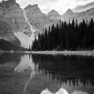 May include: A black and white photograph of a mountain lake with a forest of pine trees reflected in the water.
