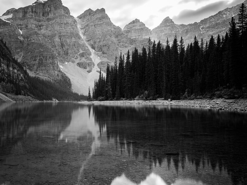 May include: A black and white photograph of a mountain lake with a forest of pine trees reflected in the water.