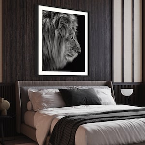 May include: Black and white photograph of a lion's head in profile, framed in a black frame and hanging on a dark wood wall above a bed with white bedding and a grey throw blanket.