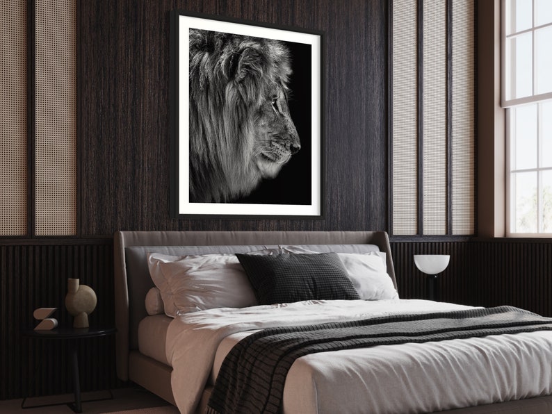 May include: Black and white photograph of a lion's head in profile, framed in a black frame and hanging on a dark wood wall above a bed with white bedding and a grey throw blanket.
