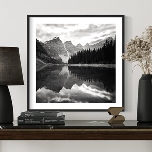 May include: Black and white photograph of a mountain lake with snow-capped mountains reflected in the water. The photo is framed in a black frame and hanging on a white wall.