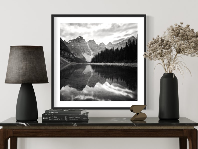 May include: Black and white photograph of a mountain lake with snow-capped mountains reflected in the water. The photo is framed in a black frame and hanging on a white wall.