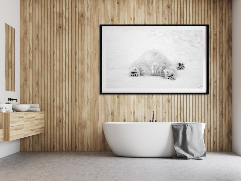 May include: A black and white photograph of a polar bear lying on its back in the snow. The photograph is framed in a black frame and hanging on a wooden wall in a bathroom.