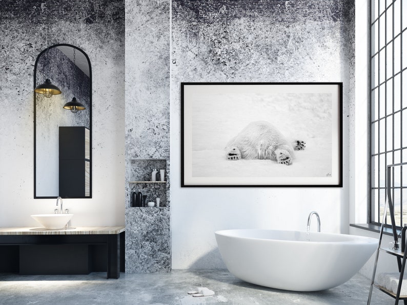 May include: A black and white photograph of a polar bear lying on its back in the snow. The photograph is framed in a black frame and is hanging on a white wall in a bathroom.