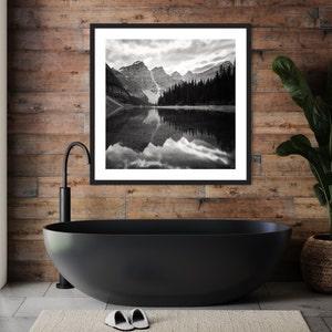 May include: Black and white photograph of a mountain lake with a reflection of the mountains in the water. The photo is framed in a black frame and hanging on a wood wall.