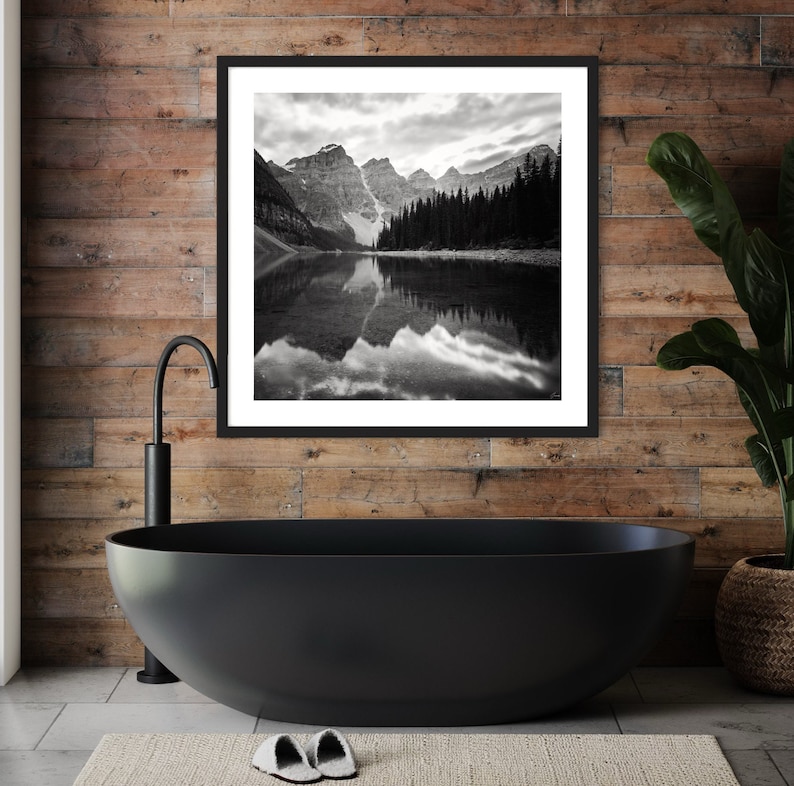 May include: Black and white photograph of a mountain lake with a reflection of the mountains in the water. The photo is framed in a black frame and hanging on a wood wall.