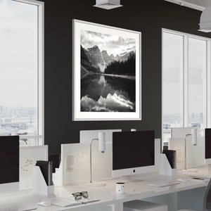May include: A black and white photograph of a mountain lake with a reflection of the mountains in the water. The photograph is framed in a white frame and is hanging on a black wall in an office setting.