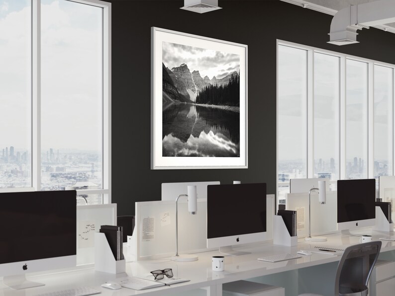 May include: A black and white photograph of a mountain lake with a reflection of the mountains in the water. The photograph is framed in a white frame and is hanging on a black wall in an office setting.