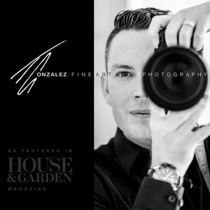 May include: Black and white portrait of a photographer holding a camera to their eye. The photographer is wearing a black shirt and a watch. The text "GONZALEZ FINE ART PHOTOGRAPHY" is visible in the image. The text "AS FEATURED IN HOUSE & GARDEN MAGAZINE" is also visible in the image.