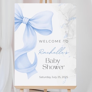 Op de afbeelding: Een welkomstbord voor een babyshower op een ezel. Het bord toont een grote, lichtblauwe strik en de woorden "Welkom bij Rachelle's Baby Shower" in elegant schrift. De datum, zaterdag 25 juli 2025, wordt ook weergegeven.