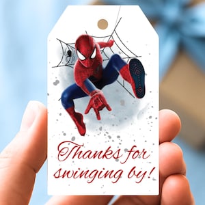 以下が含まれることがあります： スパイダーマンがダイナミックなポーズをとるイラストが描かれた白いギフトタグ。クモの巣のデザインが背景にあります。「Thanks for swinging by!」のテキストが赤いスクリプトで書かれています。ギフトに付けるための小さな穴が上部にあります。