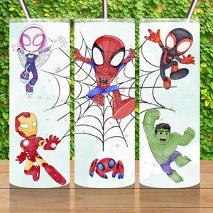 May include: Three white stainless steel tumblers with superhero designs. The tumblers feature illustrations of Spider-Man, Iron Man, Hulk, and Spider-Gwen. Each tumbler has a silver straw and is displayed on a wooden surface.