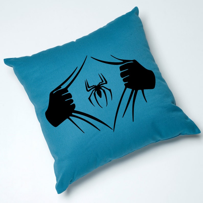 Puede incluir: Una almohada de color azul turquesa con una silueta negra de una ara&ntilde;a que sale de la tela. La ara&ntilde;a est&aacute; en el centro de la almohada.