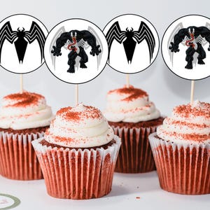 May include: Four red velvet cupcakes with white frosting and red sprinkles topped with round paper cupcake toppers featuring a black and white cartoon image of Venom from the Marvel comics.
