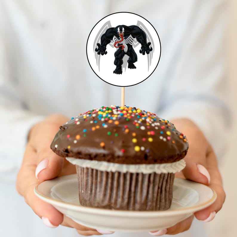 May include: A chocolate cupcake with colorful sprinkles topped with a round cake topper featuring a black and white illustration of Venom, a Marvel Comics superhero.