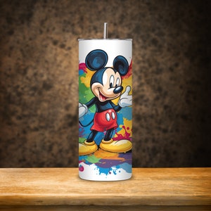 May include: A white stainless steel tumbler with a colourful paint splatter design featuring Mickey Mouse. Mickey Mouse is wearing red shorts and yellow shoes.