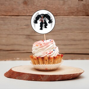 May include: A cupcake with pink and white frosting and colorful sprinkles topped with a round white cake topper featuring a black and white illustration of Venom, a Marvel Comics superhero.