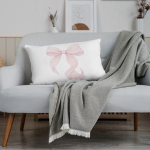 Peut inclure: Un coussin rectangulaire blanc orn&eacute; d'un motif de n&oelig;ud rose. Il est pos&eacute; sur un canap&eacute; gris clair, avec une couverture grise tiss&eacute;e. La sc&egrave;ne se d&eacute;roule dans une pi&egrave;ce lumineuse, agr&eacute;ment&eacute;e d'&eacute;l&eacute;ments d&eacute;coratifs.