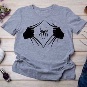 Puede incluir: Una camiseta gris con un gr&aacute;fico negro de una ara&ntilde;a que sale de la camiseta. La ara&ntilde;a est&aacute; en el centro del gr&aacute;fico y tiene ocho patas.