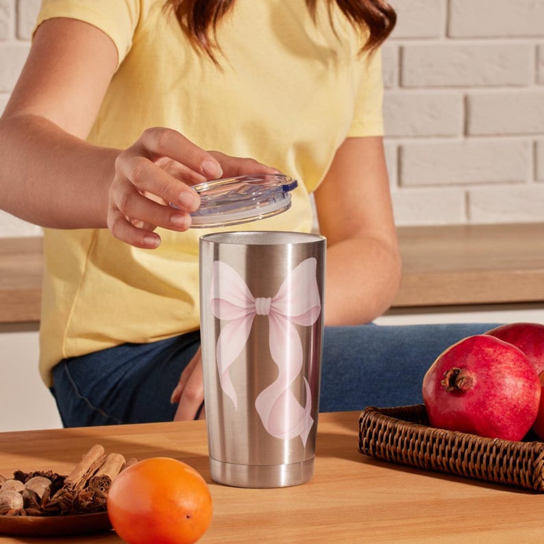 Peut inclure: Un gobelet en acier inoxydable avec un couvercle transparent. Le gobelet pr&eacute;sente un grand motif de n&oelig;ud rose clair. L'arri&egrave;re-plan comprend une surface en bois, un t-shirt jaune et un panier de grenades.