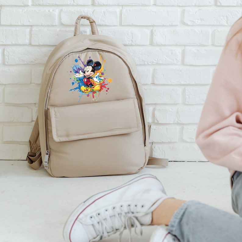 May include: A beige backpack with a colorful Mickey Mouse graphic on the front. The backpack has a large front pocket and a zipper closure.