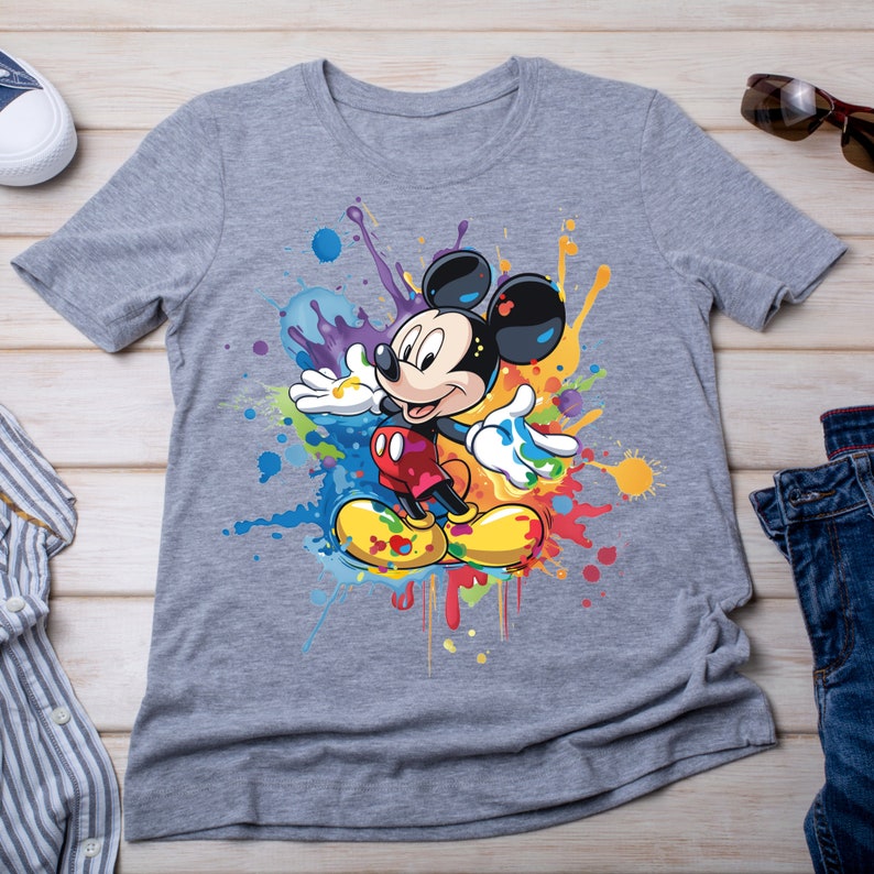 May include: A gray t-shirt with a colorful paint splatter design featuring Mickey Mouse in the center. Mickey Mouse is wearing red shorts and yellow shoes.