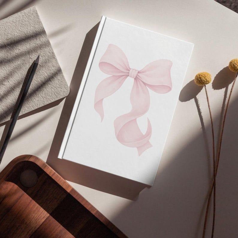 Peut inclure: Un journal blanc avec une illustration &agrave; l'aquarelle d'un n&oelig;ud rose. Le n&oelig;ud a des rubans fluides et est centr&eacute; sur la couverture. Le journal est sur une surface neutre avec un crayon, un bloc-notes et un plateau en bois.