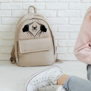 Puede incluir: Una mochila beige con un gr&aacute;fico de ara&ntilde;a negra en la parte delantera. La ara&ntilde;a se muestra saliendo de la mochila, como si fuera un superh&eacute;roe.