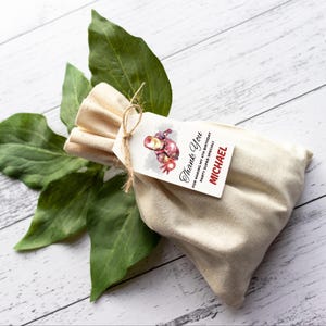 May include: A beige fabric gift bag with a tag tied with twine. The tag features an Iron Man illustration and the text "Thank You" and "MICHAEL". Green leaves are in the background.