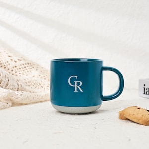 Pode incluir: Caneca de cerâmica azul-turquesa com o logotipo branco « GR ». A caneca tem uma base cinza e uma alça curva. Um pequeno biscoito repousa sobre a superfície ao lado da caneca. O fundo é branco neutro.