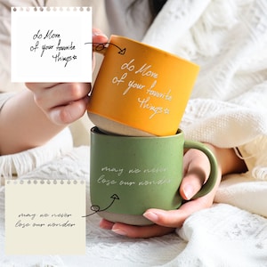 Peut inclure: Deux tasses en céramique, une orange et une verte, empilées. La tasse orange porte l'inscription "do More of your favorite Things" et la tasse verte porte l'inscription "may we never lose our wonder". Les deux tasses ont une base de couleur neutre.