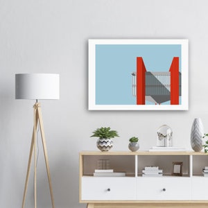 May include: A framed print of a minimalist illustration of a red building with a light blue sky background. The building has a geometric design with a flat roof and large windows. The print is displayed on a shelf with a lamp, plants, and other decorative items.