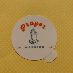 Puede incluir: Pegatina redonda blanca con una ilustración azul y naranja de manos en oración. El texto "Prayer Warrior" está escrito en azul y naranja debajo de las manos.