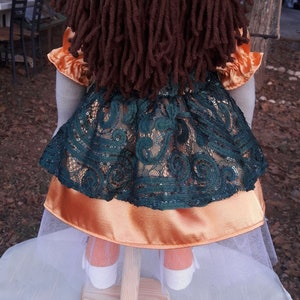 Puede incluir: Una mu&ntilde;eca hecha a mano con pelo largo de lana marr&oacute;n, que lleva un vestido de encaje verde con una falda interior de sat&eacute;n naranja y una falda de tul blanca.