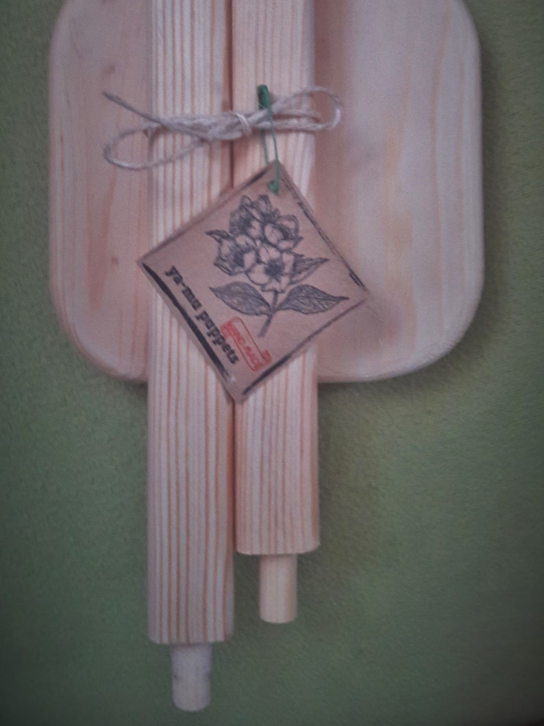 Puede incluir: Dos clavijas de madera atadas juntas con cuerda y una peque&ntilde;a etiqueta que dice "Je m'appelle" con un dise&ntilde;o floral. Las clavijas est&aacute;n sobre una superficie de madera.