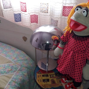 Puede incluir: Un vestido de lunares rojos y negros con cuello blanco es usado por un t&iacute;tere con cabello de lana rubio y una boca grande abierta. El t&iacute;tere est&aacute; sentado en una mesa con una l&aacute;mpara y una colcha estampada de colores en el fondo.