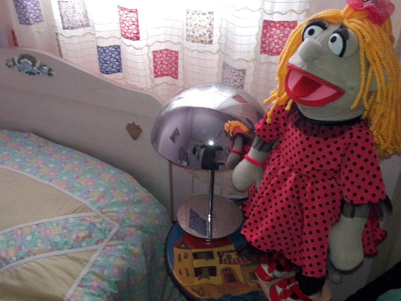 Puede incluir: Un vestido de lunares rojos y negros con cuello blanco es usado por un t&iacute;tere con cabello de lana rubio y una boca grande abierta. El t&iacute;tere est&aacute; sentado en una mesa con una l&aacute;mpara y una colcha estampada de colores en el fondo.