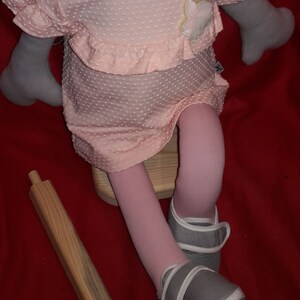 Puede incluir: Un vestido rosa con lunares blancos y una aplicaci&oacute;n de flores. El vestido lo lleva una mu&ntilde;eca con medias rosas y zapatos grises. La mu&ntilde;eca est&aacute; sentada en un taburete de madera.