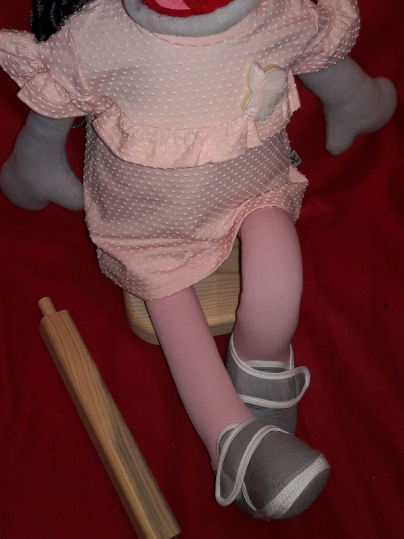 Puede incluir: Un vestido rosa con lunares blancos y una aplicaci&oacute;n de flores. El vestido lo lleva una mu&ntilde;eca con medias rosas y zapatos grises. La mu&ntilde;eca est&aacute; sentada en un taburete de madera.