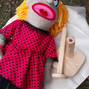 Puede incluir: Un t&iacute;tere gris con pelo amarillo que lleva un vestido rojo con lunares negros. El t&iacute;tere est&aacute; tumbado sobre una superficie blanca con un soporte de madera.