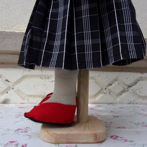 Puede incluir: Una mu&ntilde;eca con una falda de cuadros negros y blancos y zapatos rojos. La mu&ntilde;eca est&aacute; de pie sobre un soporte de madera.