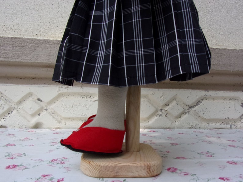 Puede incluir: Una mu&ntilde;eca con una falda de cuadros negros y blancos y zapatos rojos. La mu&ntilde;eca est&aacute; de pie sobre un soporte de madera.