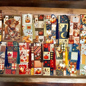 May include: A collection of rectangular paper bookmarks featuring various autumn-themed patchwork patterns, including illustrations of pumpkins, sunflowers, cats, geese, and plaid designs. Several bookmarks are accented with colorful fabric tassels.