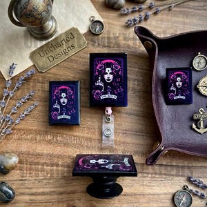 May include: A collection of accessories featuring a gothic tarot card design with a woman, roses, and the text "The Bitch." Items include a badge reel, a phone grip, and a small pin. The background includes a globe and compass.