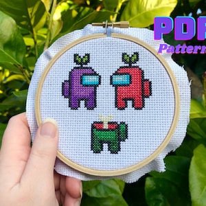 Puede incluir: Un aro de bordado blanco con un patrón de punto de cruz de tres personajes de Among Us. Los personajes son morado, rojo y verde. Los personajes tienen una planta en la cabeza. El aro está en la mano de una persona.