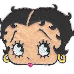 May include: Embroidered patch featuring the face of a cartoon character with black hair, light skin, and large blue eyes. The character has a small pink mouth and yellow earrings. The patch is likely for clothing or accessories.